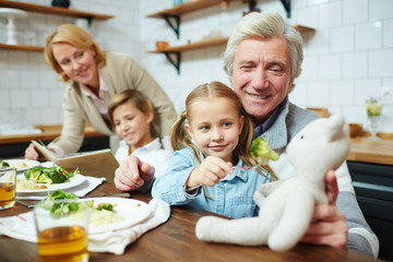 Happy mature man holding teddybear while his granddaughter giving food to her toy by dinner