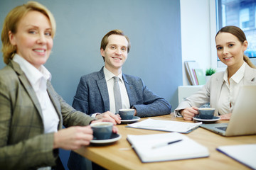 Team of successful leaders sitting by workplace in office and looking at you