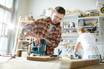 Young woman processing wooden board with electric fretsaw in working environment