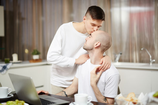Amorous young man kissing his networking sweetheart on head at home