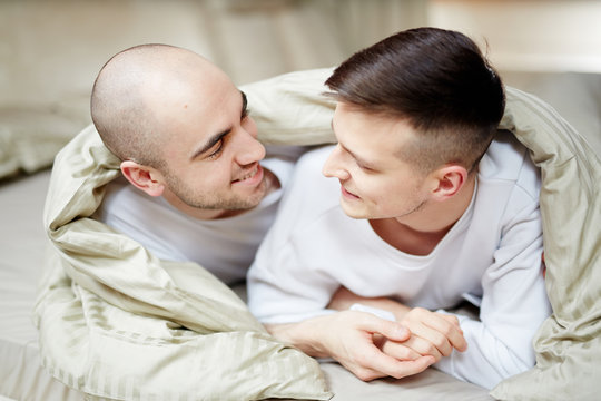Two amorous guys lying on bed under blanket and lookign at one another with smiles