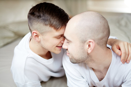 Two young gay lovers in white T-shirts touching by their heads in bed