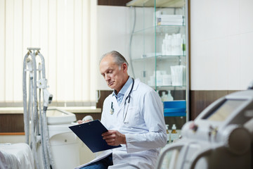 Senior doctor with clipboard making notes in medical card of one of his patients