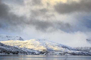 Sunset at the lakeside with rocks of a fjord during a snow storm in a snowy winter landscape.