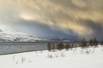 Fototapeta na wymiar Sunset at the lakeside with rocks of a fjord during a snow storm in a snowy winter landscape.