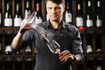 Bartender pours red wine in glass from big transparent vessel