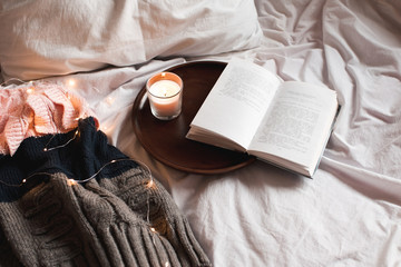 Open book with burning candle on wooden tray, knitted sweater in bed. Good morning. Cozy atmosphere.