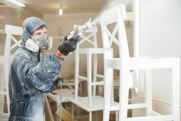 Man painting chair into white paint in respiratory mask. Application of flame retardant ensuring fire protection, airless spraying.