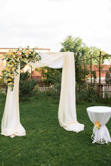 Open air decorated area for the wedding ceremony with a wooden arch decorated with fresh flowers and beige material. Beautiful wedding set up. Wedding ceremony on green lawn in the garden. Registratio