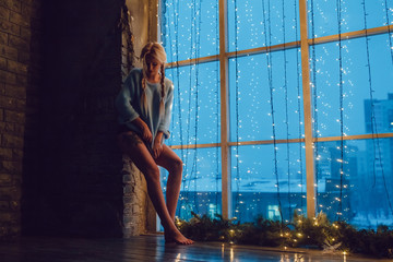 Naklejka na ściany i meble Sexy blond girl in a sweater stands near a window.