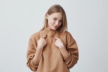 Shot of beautiful attractive blonde woman smiling, pointing with index fingers at herself, dressed in beige long-sleeved sweater, expressing positive emotions and feelings. Beauty and youth concept