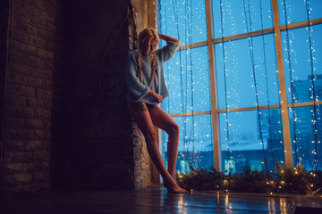Sexy blond girl in a sweater stands near a window.