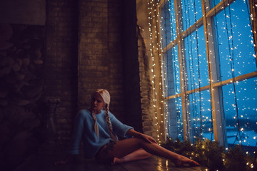 Sexy blond girl in a sweater sits near a window with lights.