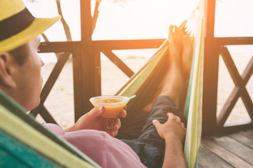 Handsome young man holding an orange cocktail and lying in a hammock at a sunny beach by an ocean