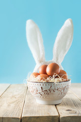 Chicken eggs in bowl with easter bunny ears on blue background