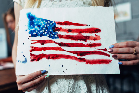 Close-up view of female hands holding a sheet of paper with a hand-darwn watercolor illustration of the flag of the United States