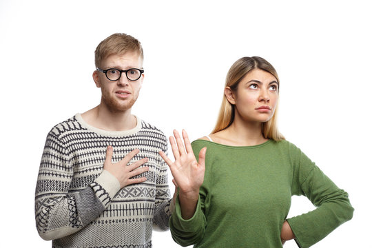 Studio shot of displeased blonde girl showing stop sign with her palm, meaning It's over while her unhappy miserable boyfriend with unhappy look holding hand on his chest as sign of broken heard