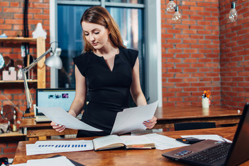 Serious woman reading papers studying resumes standing at work desk in stylish office