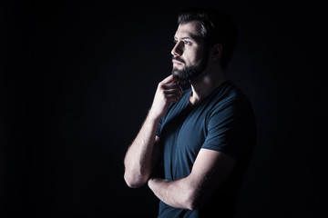 Involved in thoughts. Serious nice brutal man holding his chin and being thoughtful while standing against black background