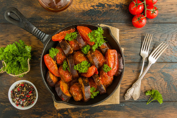 Black cast-iron skillet with Turkish kebab cooked with eggplant and tomatoes on rustic wooden table, top view.