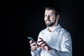 New smartphone. Handsome positive pleasant man holding a modern gadget and smiling while standing against black background