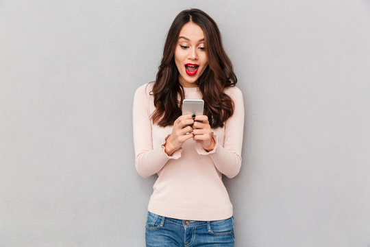 Image of gorgeous woman in casual being surprised or excited to receive pleasant text message on her cell phone over gray background