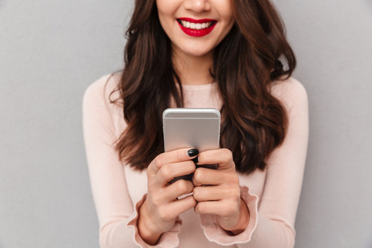 Close up image of young smiling female with red lips holding mobile phone chatting or playing games via modern device over gray background cropped