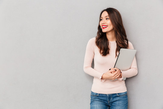 Image of cute female with beautiful smile holding silver notebook looking aside over grey wall