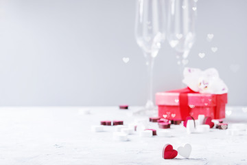 Love Valentine day greeting card with two empty champagne glasses, red gift box with orchid flower and wooden hearts over white gray background. Copy space. Selective focus