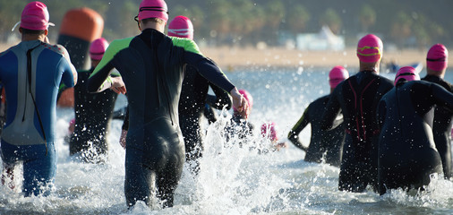 Group triathlon participants running into the water for swim portion of race,splash of water and athletes running - obrazy, fototapety, plakaty