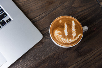Latte art cup on wooden table with keyboard of laptop in modern office