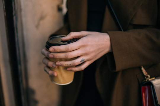 Fashionable woman in coat, stands outdoors on street, holds in hand with tattooed fingers, take away cup with coffee to go. Vintage look and feel nostalgia photo, tender and sensual morning routine
