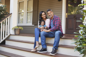 Family With Baby Son Sit On Steps Leading Up To Porch Of Home