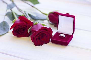 The concept of Love, Wedding, Proposal, Anniversary, St. Valentine's Day, Mother's Day with beautiful red roses and a wedding ring in a red velvet box in a close up, light wooden background