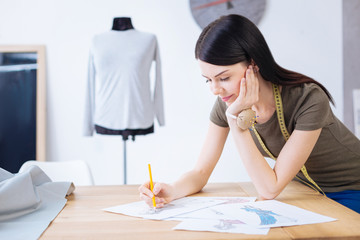 Calmness. Creative talented young worker of a professional atelier sitting with her chin resting on her hand and drawing a sketch of a future dress