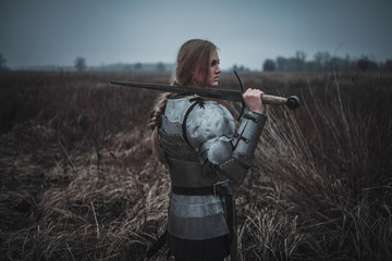 Girl in image of Jeanne d'Arc in armor and with sword in her hands stands on meadow. Back view.