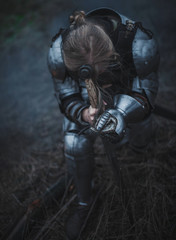 Girl in image of Jeanne d'Arc in armor and with sword in her hands kneels against background of dry...