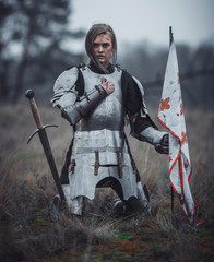 Girl in image of Jeanne d'Arc in armor kneels with flag in her hands and sword on meadow.