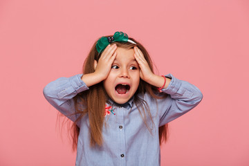 Thrilled little girl reacting emotionally grabbing head with both hands being shocked screaming over pink background - obrazy, fototapety, plakaty