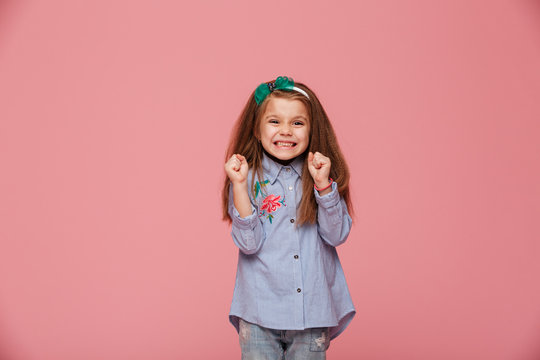 Smiling girl model in hair hoop and fashion clothes expressing happiness gesturing with clenched fists against pink background