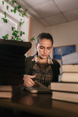 a beautiful slim girl in a knitted green cardigan with a dark hair holds her hand near the chin and sees at right near piles of large books in the library