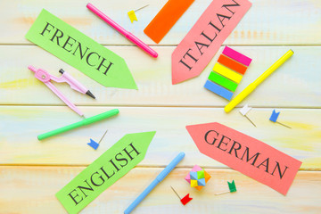 Learning Foreign Languages concept: paper arrows French, English, Italian, German and bright school supplies on a light colored wooden background, close up, top view