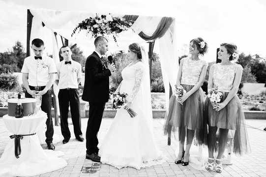 Wedding couple making vows outdoor under the floral arch in front of many guests. Black and white photo.