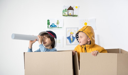 Two young boys standing inside of carton boxes, one of them is looking aside through rolled piece of paper. Childish drawings hanging on background