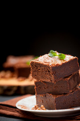 Chocolate brownie square pieces in stack on white plate with walnuts, decorated with mint leaves and cocoa on black background. Delicious dessert. Dark mood. Close up photography. Selective focus