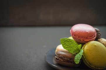 Assortment of color macaroon cookies on plate, decorated with mint leafs, dark background. green pistachios, brown chocolate, pink strawberry and creme vanilla almond cakes. copyspace for text