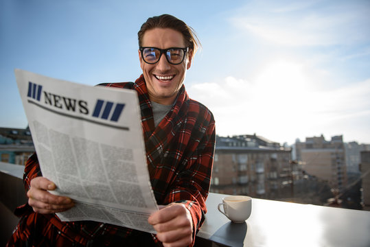 Portrai of cheerful man looking at camera with radiant smile. He is standing on the balcony and holding the newspaper. Copy space in right side