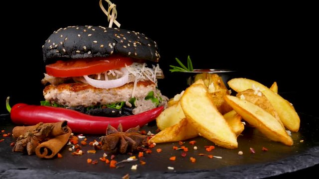 Big black burger with Fried Potato wedges and tomatoes sauce, decorated with pepper corns and chili pepper, on black stone tray, loop, 4K, 60 FPS