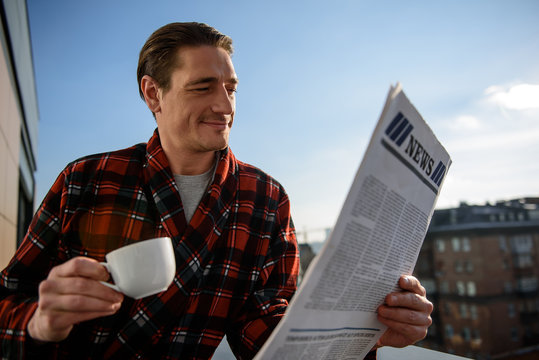 Waist up portrait of satisfied guy reading the newspaper with calm look. He is standing on terrace and holding a cup in his hand