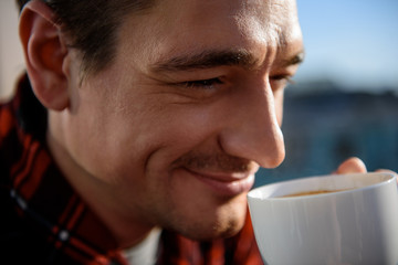 Close up portrait of happy adult man drinking coffee outside in the morning. He is holding a cup in hand with calm look. Focus on his face